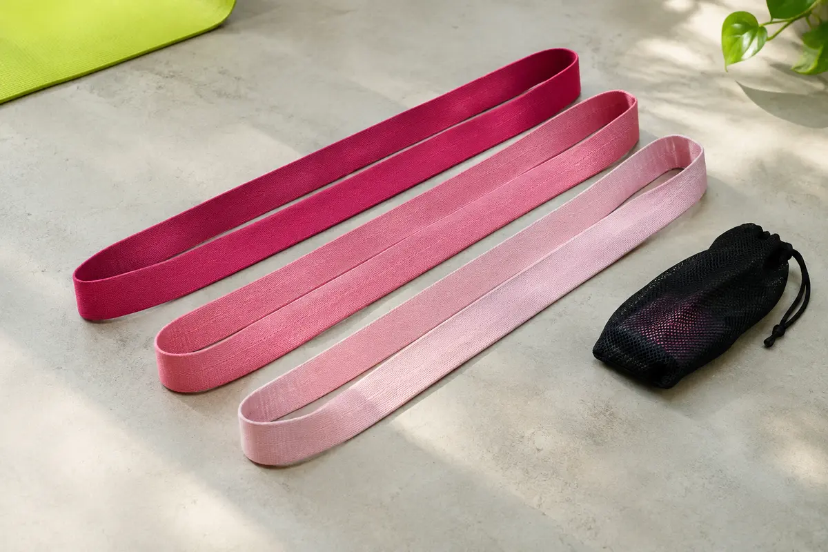 Trois longues bandes de résistance en tissu rose, étendues côte à côte près de leur sac en maille.