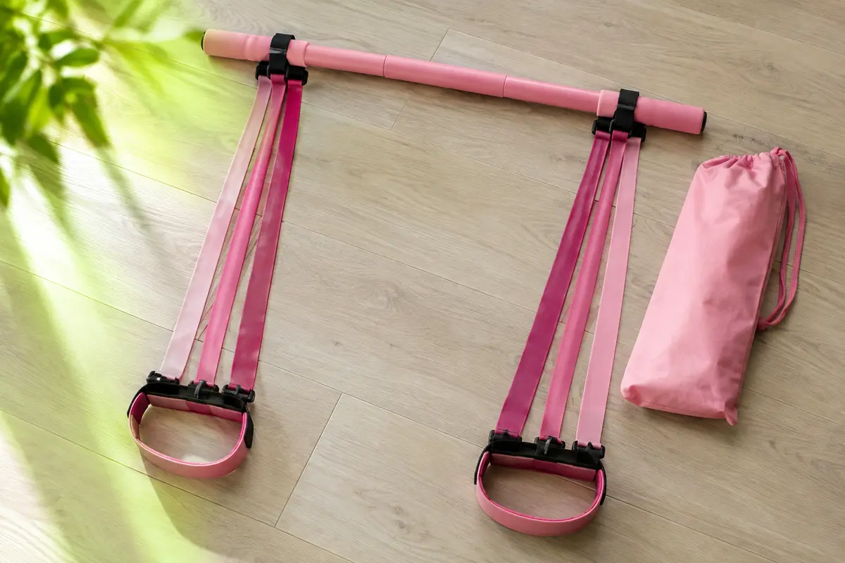 Barre de Pilates rose couchée sur un parquet, reliée par six bandes roses empilées à deux pédales en sangle, avec un sac de rangement.