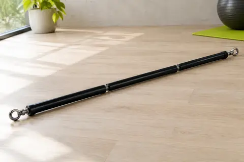 Barre de Pilates noire démontable en trois parties, avec œillets métalliques pivotants, posée sur un sol clair.