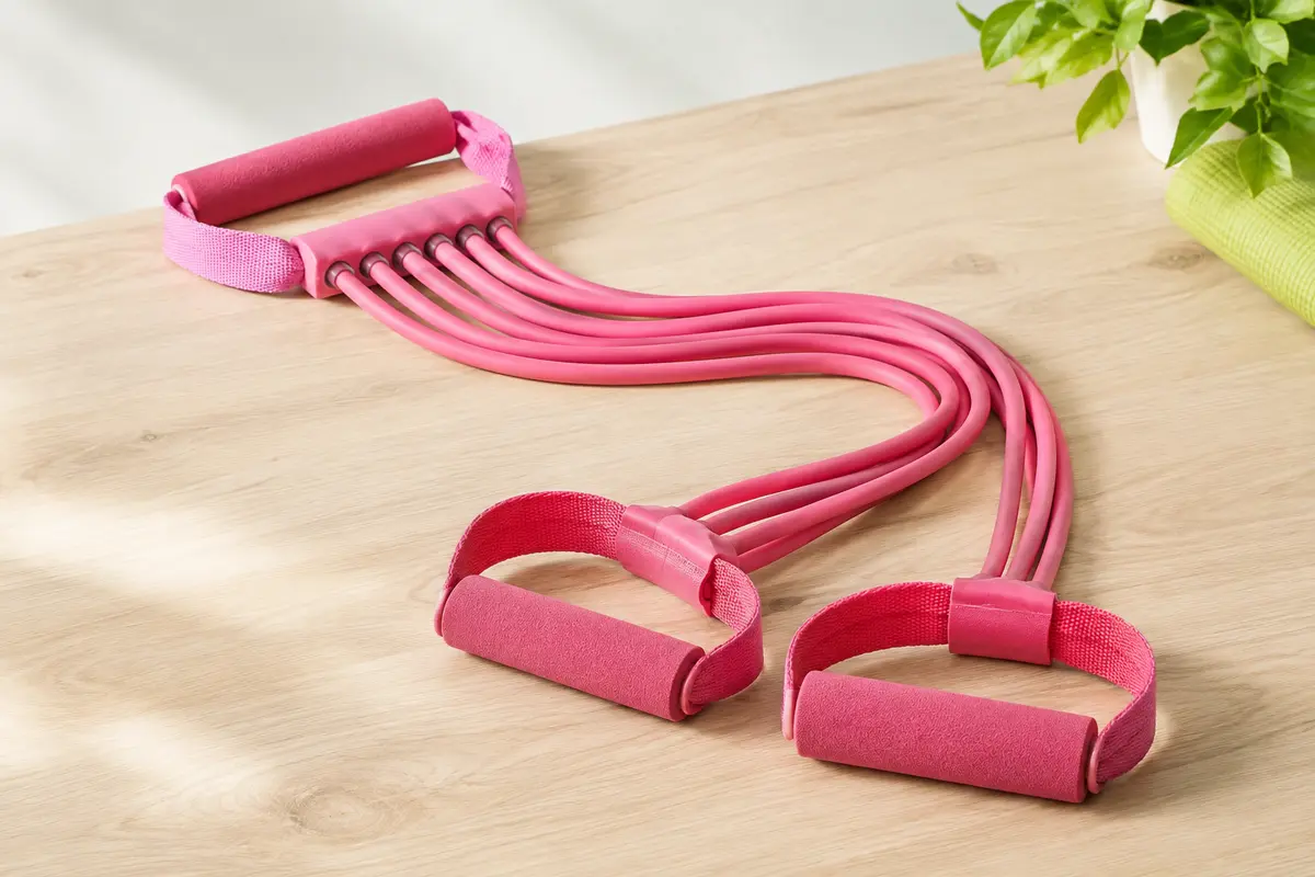 Élastique de fitness rose à six tubes avec poignées et pédales en mousse, posé sur une surface claire.