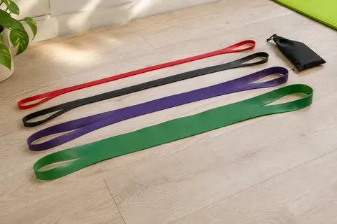 Quatre longues bandes élastiques en boucle, rouge, noire, violette et verte, étendues côte à côte sur un parquet clair.